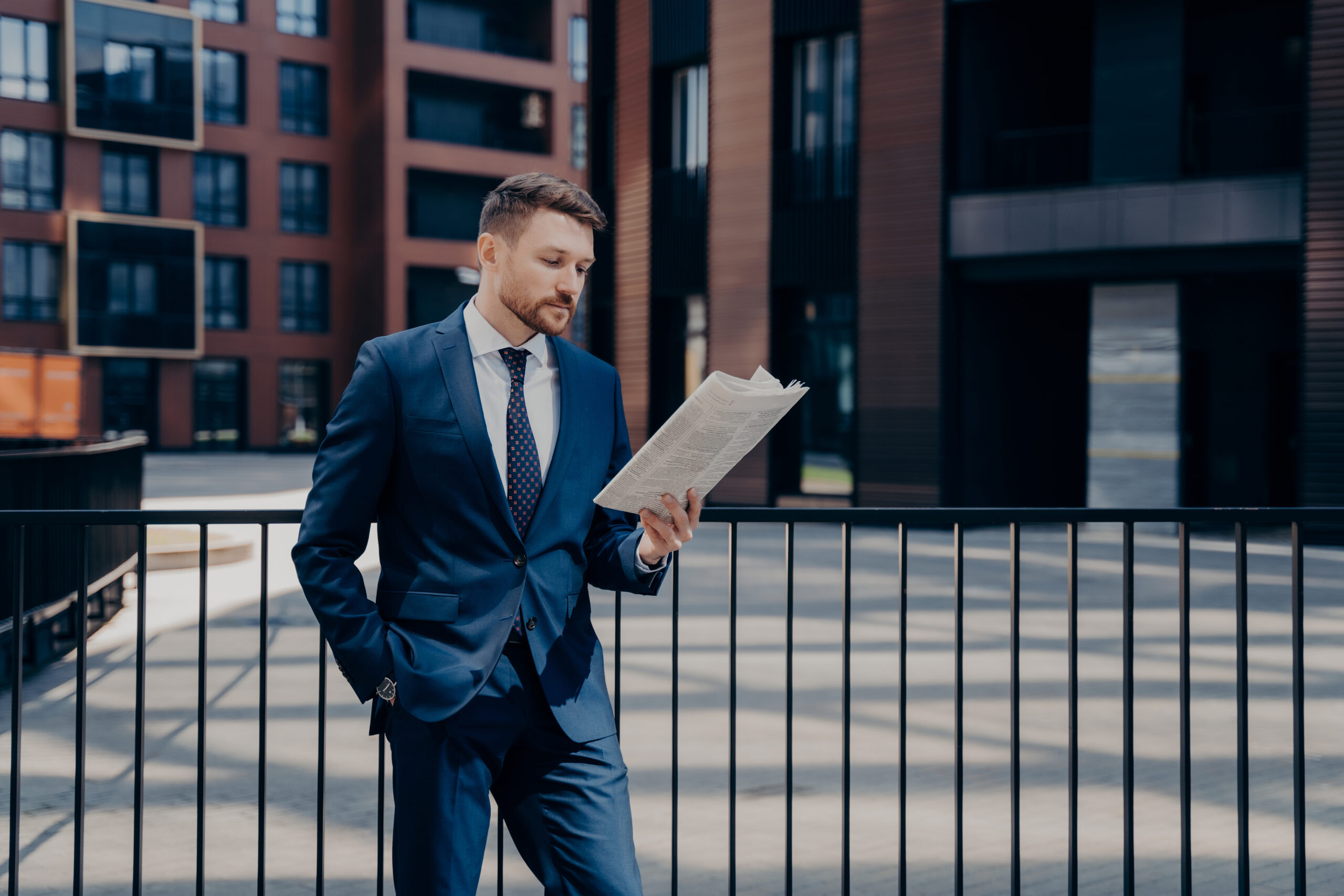 Handsome male boss wearing blue suit, reading newspaper thoughtfully, looking for news regarding his firm, standing alone with one hand in his pocket in front of corporate buildings